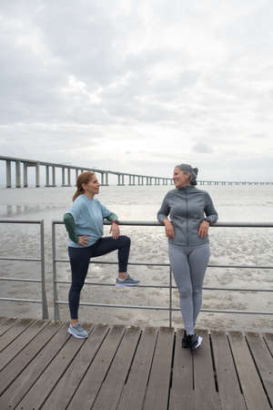 Happy senior friends resting after training. Women in sportive clothes communicating on embankment...