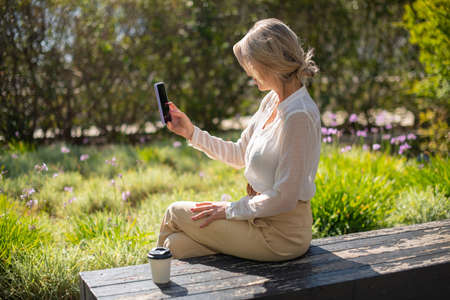 Businesswoman taking selfie outdoors. Female model in formal clothes sitting in garden. Technology,...