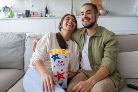 Happy couple eating popcorn and watching TV. Woman and man sitting on sofa, holding box of popcorn, laughing. Food, leisure conceptの写真素材