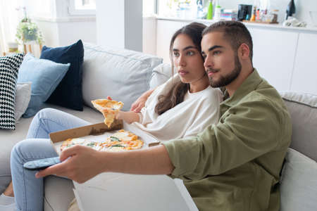 Content couple eating pizza and watching TV. Woman and man sitting on sofa, holding cheese pizza,...