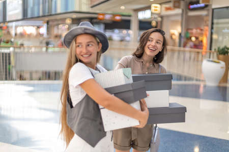 Happy young women standing with boxes at shopping mall and smiling. Stylish girls doing shopping together. Shopping and sale conceptの写真素材