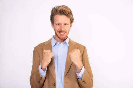 Portrait of happy young businessman standing with clenched fists. Caucasian man wearing blue shirt and beige woolen suit coat smiling at camera and making winner gesture. Success conceptの写真素材