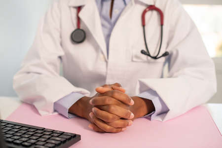 Close-up of clutched hands of female general practitioner sitting at her workplace. Young African American doctor wearing lab coat and stethoscope sitting at computer. Medical service conceptの写真素材