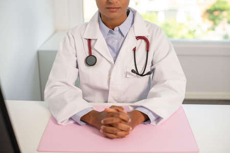 Serious young female general practitioner sitting at table with clutched hands. Close-up of African American doctor wearing lab coat and stethoscope in her office. Woman doctor conceptの写真素材