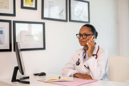 Portrait of confident female doctor answering call in office. Young African American medic talking on mobile phone at her workplace. Medical service conceptの写真素材