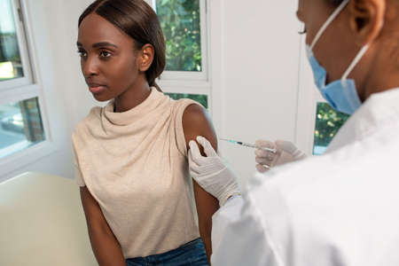 Nurse vaccinating young African American woman in clinic. Female patient sitting in doctors office...