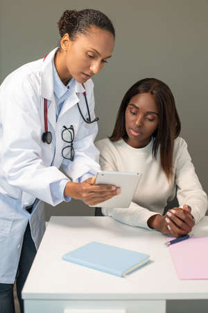 Confident doctor showing medical tests on touchpad to woman. Young African American general practitioner consulting female patient in clinic. Medical consultation conceptの写真素材