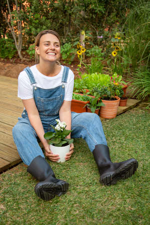 Delighted woman with potted plant. Woman in jeans overall sitting, holding flower in pot. Gardening, hobby conceptの写真素材