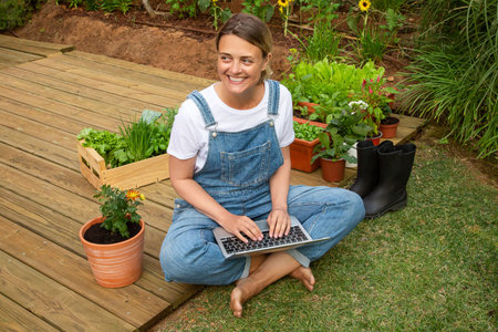 Calm woman using laptop in garden. Female gardener in overall sitting on ground. Technology, nature,...