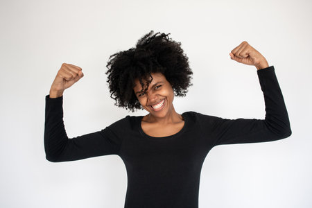 Portrait of excited African American woman showing muscles. Young female model wearing black dress...