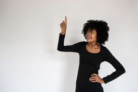 Portrait of happy African American woman pointing upwards. Young female model wearing black dress standing and looking up against white background. Advertisement conceptの写真素材