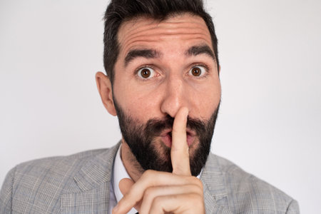Handsome man with beard calling be silent. Male model in suit touching lips with finger, looking at camera. Portrait, studio shot, gesture conceptの写真素材