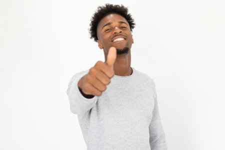 Close-up of happy African American man showing thumb up. Young bearded guy wearing white sweater stretching arm with like gesture and smiling at camera. Success and approval conceptの写真素材