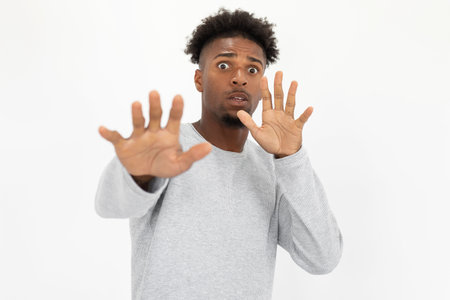 Portrait of terrified African American man making stop gesture. Young bearded guy wearing white sweater looking at camera in horror against white background. Horror and terror conceptの写真素材