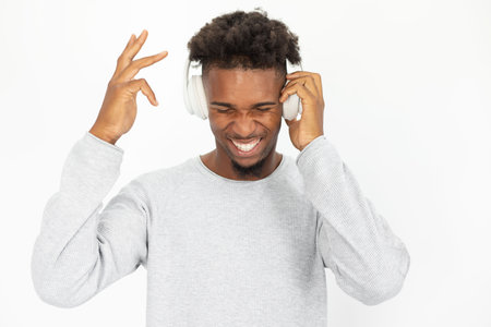 Excited African American man listening to music in headphones. Young bearded guy wearing white...