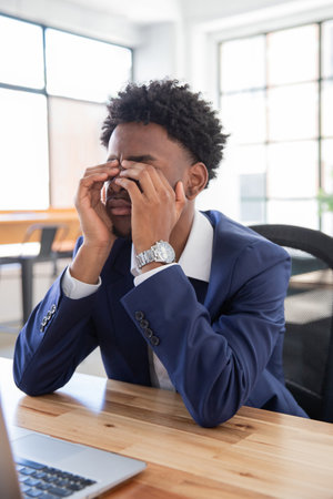 Portrait of unhappy male office worker rubbing eyes. Tired businessman sitting at desk, covering...
