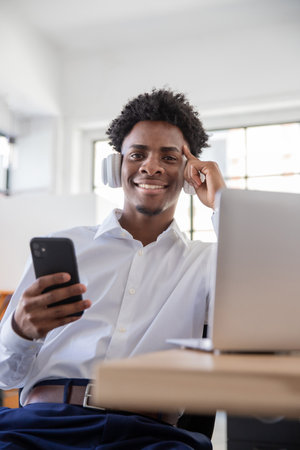 Portrait of happy male office worker in headphones using phone. Smiling businessman looking at...