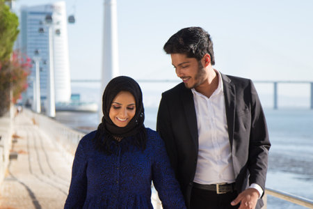 Close-up of happy man and woman walking together. Young Muslim couple walking along summer city quay, smiling man in black suit looking with love at woman in colored dress. Dating and romance conceptの写真素材