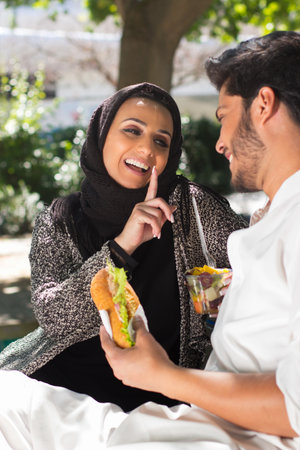 Portrait of young Arabian couple resting in park. Happy smiling man with burger and beautiful woman...