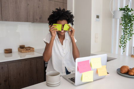Happy businesswoman sitting at desk with stickers on her eyes. Smiling Black woman having fun while working from home. Game, home office, enjoyment conceptの写真素材