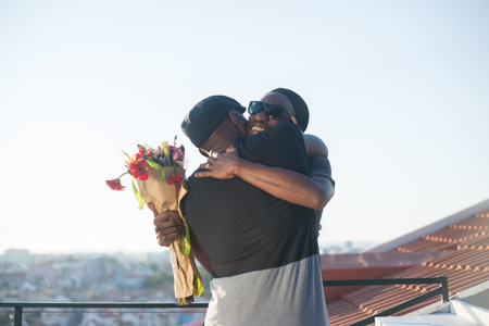 Portrait of African American gay men embracing on roof top. Two happy men standing on blue sky background hugging each other one man holding bunch of flowers. LGBT couples relations, equality conceptの写真素材