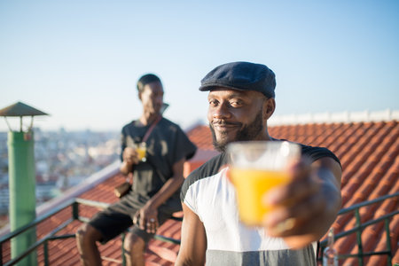 Close-up of happy African man holding glass of fruit juice. Young bearded man in black cap standing on roof top smiling stretching hand with glass of juice to camera. Rest and human relations conceptの写真素材