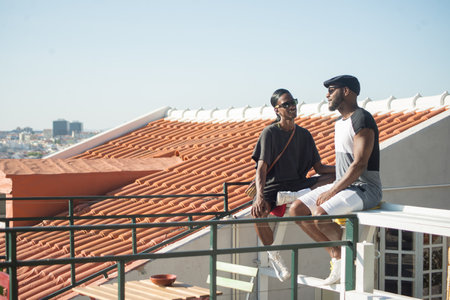 Portrait of Afro American gay men communicating. Two happy boyfriends dating on roof top sitting together on bench talking on blue sky and summer city background. LGBT couples love, relations conceptの写真素材