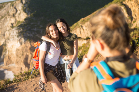 Woman taking photo of teen daughters against rock. Sisters embracing, looking at camera and smiling...