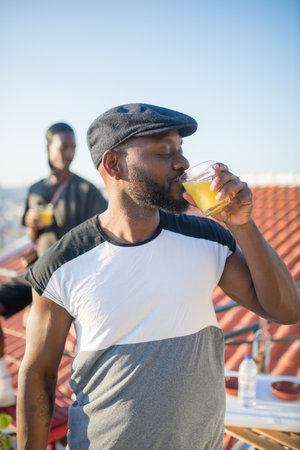 Close-up of African American man drinking juice on roof top. Handsome man in black cap standing drinking juice and his friend sitting on fence behind him.Spending free time, leisure, relations conceptの写真素材