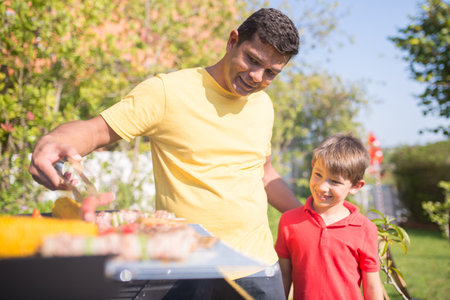 Happy father grilling meat and vegetables with son. Dark-haired man in yellow T-shirt putting sausage on BBQ grid. Smiling little boy standing next to him. BBQ, cooking, food, family conceptの写真素材