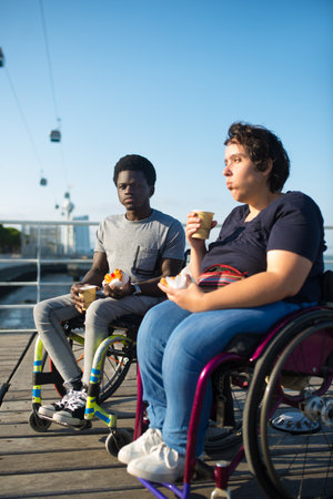 Content biracial couple drinking coffee on sunny day. African American man and Caucasian woman in wheelchairs on embankment, drinking hot beverage from cups. Snack, relationship, happiness conceptの写真素材