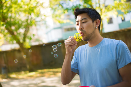 Close-up of young daddy blowing soap bubbles for his kid in park. Happy Asian man in casual clothes holding bottle with soap solution and showing how to blow bubbles. Vacation and parenthood conceptの写真素材
