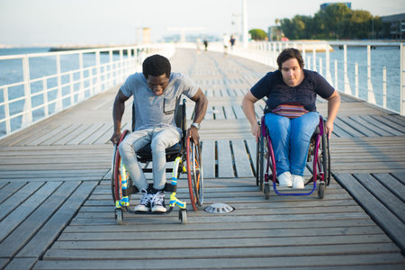 Outgoing multicultural couple spending day together. African American man and Caucasian woman in wheelchairs on embankment, racing against each other. Love, affection, happiness conceptの写真素材