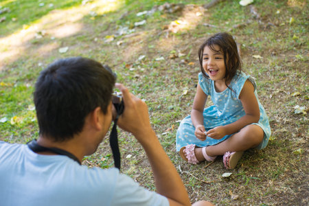 Portrait of little Asian girl and father taking photo of her. Beautiful girl in summer dress sitting...