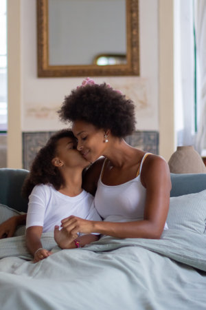 Tender mother and daughter with curly hair. African American woman and little girl sitting in bed in morning, child kissing parent on cheek. Family, parenthood, home conceptの写真素材