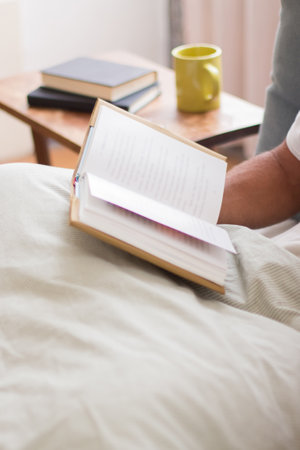 Unrecognizable man lying on bed in morning and holding book. Man reading novel after waking up, enjoying time alone, devoting time to hobby. Morning routine, literature, sleep conceptの写真素材