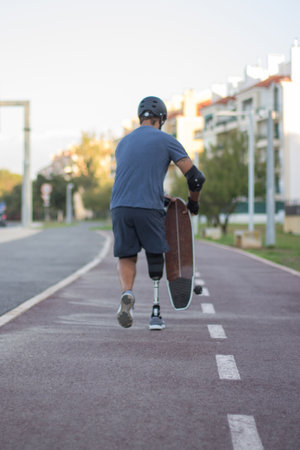 Back view of man with mechanical leg in casual clothes. Mid adult sportsman running down special road, holding skateboard. Sport, disability, training conceptの写真素材