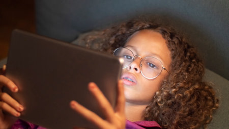 Portrait of cute girl using digital gadget. Curly little girl resting in bed and watching her tablet...