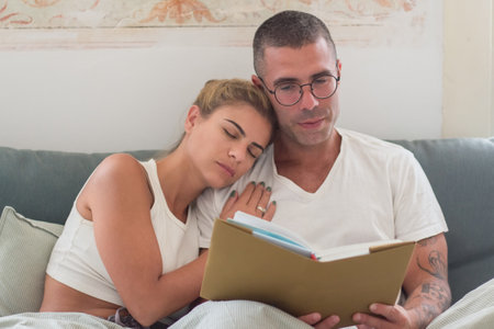 Man reading book in bed while his wife sleeping on his shoulder. Focused Caucasian man in eyeglasses and pajamas reading novel in early morning, devoting time to hobby. Love, sleep cycle conceptの写真素材