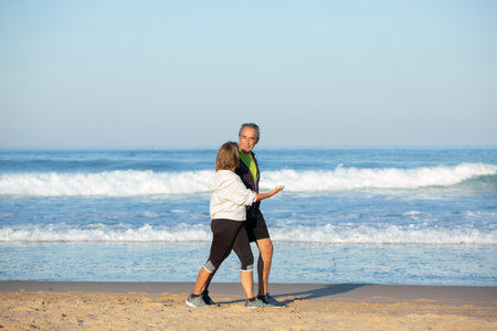 Cheerful senior couple talking while walking along shore. Long shot of beautiful mature man and woman in sportswear spending time together, looking at each other with tenderness. Communication conceptの写真素材