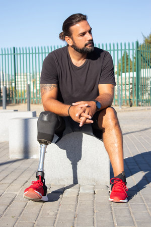 Portrait of man with disability sitting on stone bench. Serious man with prosthetic leg resting outdoors after training sitting looking aside. Active life of people with disability and health conceptの写真素材