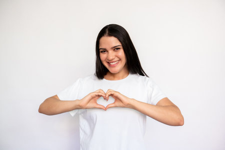 Portrait of happy young woman making heart gesture over white background. Caucasian lady wearing white T-shirt showing love or gratitude symbol. Love and admiration conceptの写真素材