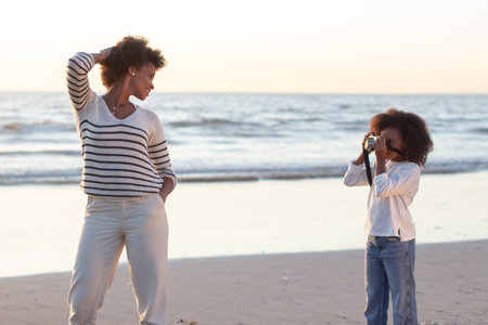 Focused little girl photographing mother on beach. Woman posing, daughter holding old-fashioned...