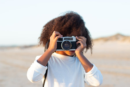 Portrait of cute girl with old-fashioned camera. Female model with curly hair and in white pullover holding camera on beach. Childhood, hobby conceptの写真素材