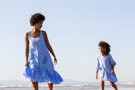 Tranquil mother and daughter on vacation. African American family in beautiful dresses walking on beach, having good time. Family, travelling, parenthood conceptの写真素材