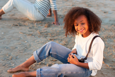 Cute girl with old-fashioned camera. Female model with curly hair and in white pullover holding camera on beach. Mother in background. Childhood, hobby conceptの写真素材