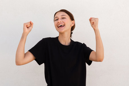 Portrait of joyful young woman celebrating success over white background. Caucasian woman wearing black T-shirt making winning gesture and laughing. Success conceptの写真素材