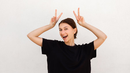 Portrait of excited young woman making bunny ears against white background. Caucasian woman wearing...