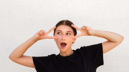 Portrait of excited young woman making victory gesture against white background. Caucasian woman wearing black T-shirt looking away with open mouth. Excitement conceptの写真素材