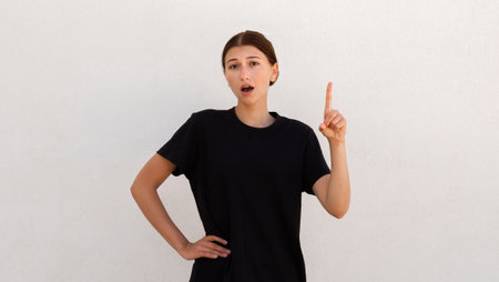 Portrait of surprised young woman pointing finger up over white background. Caucasian woman wearing black T-shirt having idea. Imagination and creativity conceptの写真素材